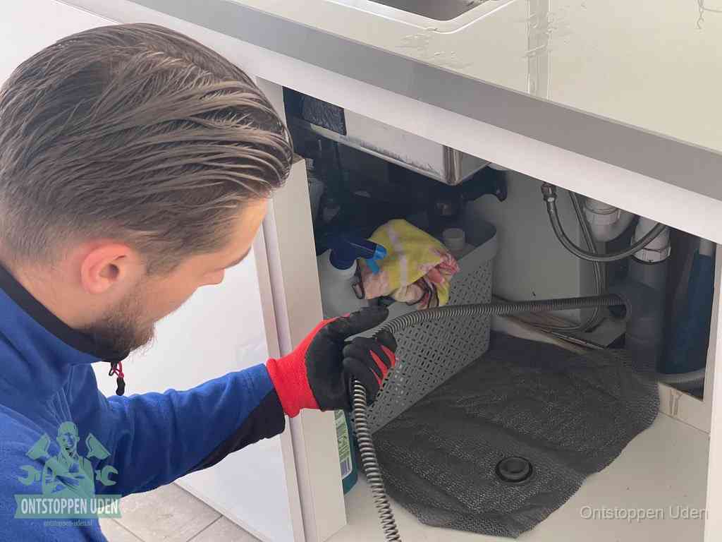 Een monteur voert een flexibele ontstoppingsveer in onder een keukenkastje om een verstopping voorbij de sifon te bereiken in Uden