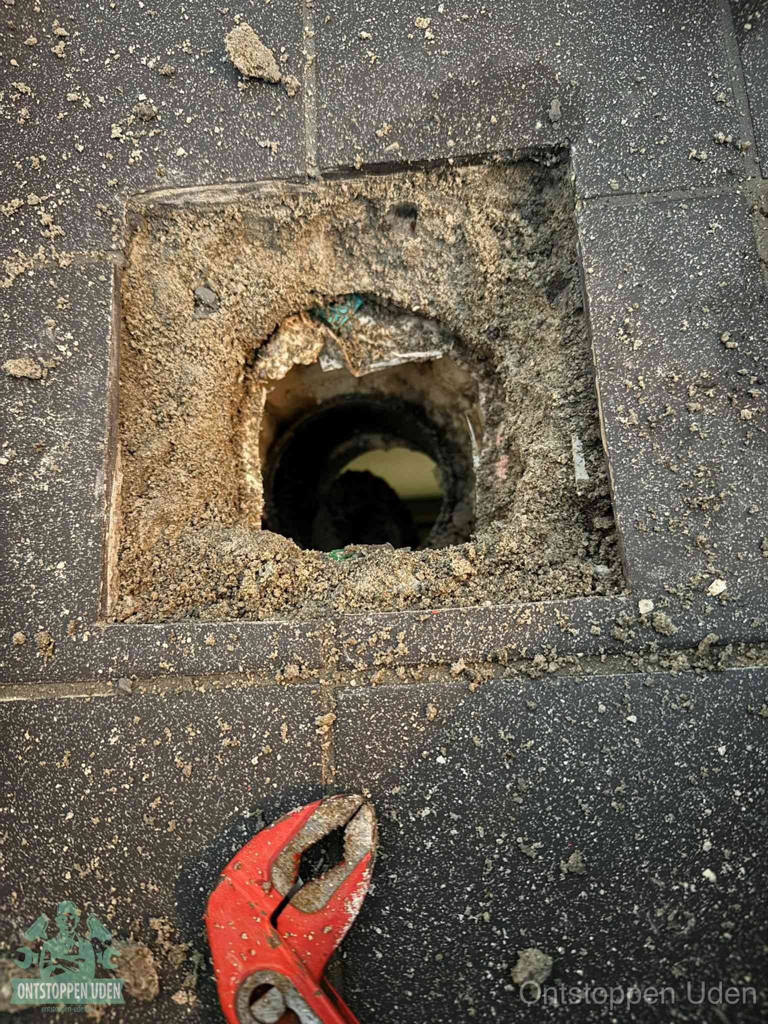 Een geopend rioolputje in een betontegelvloer met losgemaakt rooster en een zichtbare ronde afvoerbuis, het achterhalen waar de afvoerverstopping zit in Uden
