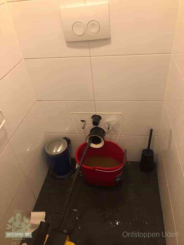 Een ontstopte wand-wc met verwijderde toiletpot waar een ontstoppingsveer in de wanduitgang verdwijnt, het achterhalen waar de verstopping zit in Uden