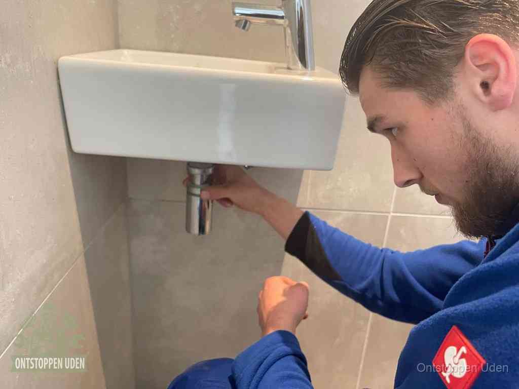Een monteur draait met de hand de zwanenhalssifon onder een fonteinwasbak in de toiletruimte los om de bron van rioollucht in Uden te vinden
