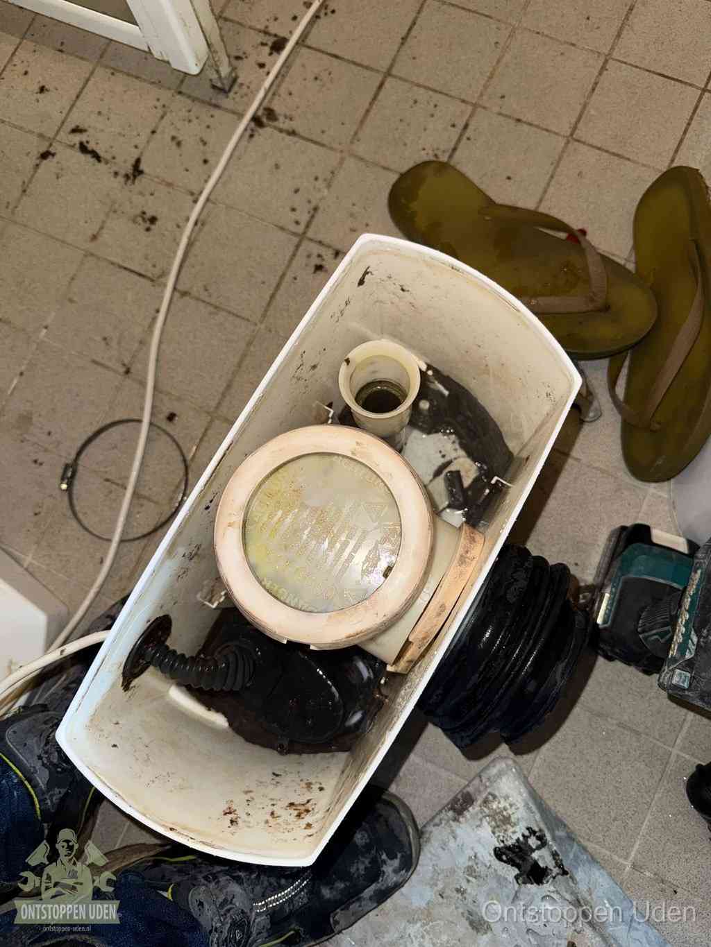 Een gedemonteerde toiletpot op de vloer met ontstoppingsveer en gereedschap ernaast, het verwijderen van een vastzittend voorwerp uit de wc in Uden